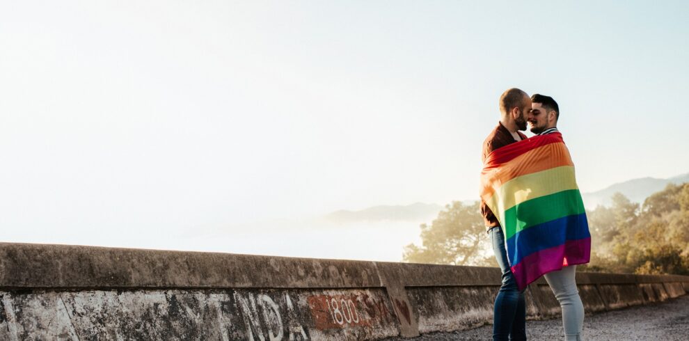 Gay couple wrapped in a gay pride flag kissing on a road in the mountains