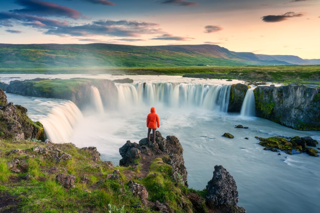 Godafoss waterfall flowing with colorful sunset sky and male tourist standing on cliff