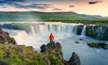 Godafoss waterfall flowing with colorful sunset sky and male tourist standing on cliff