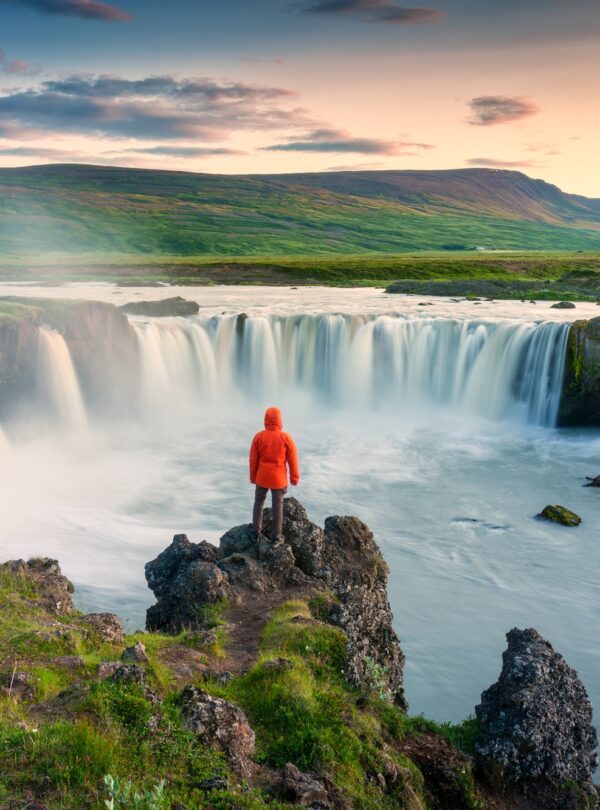 Godafoss waterfall flowing with colorful sunset sky and male tourist standing on cliff
